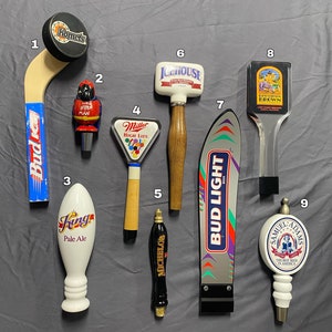 May include: A collection of beer tap handles with various designs. Includes a hockey stick-shaped handle with "Komet" and "Bud" branding, a red and yellow figure, and handles for "King Pale Ale", "Michelob", "Icehouse", "Bud Light", and "Samuel Adams".
