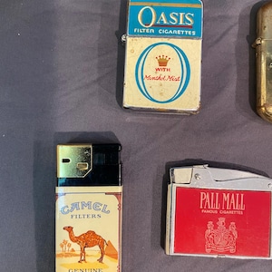 May include: Four vintage cigarette lighters with different brand names. The lighters are silver, gold, and red with various designs. The lighters are all in good condition and have a retro look.
