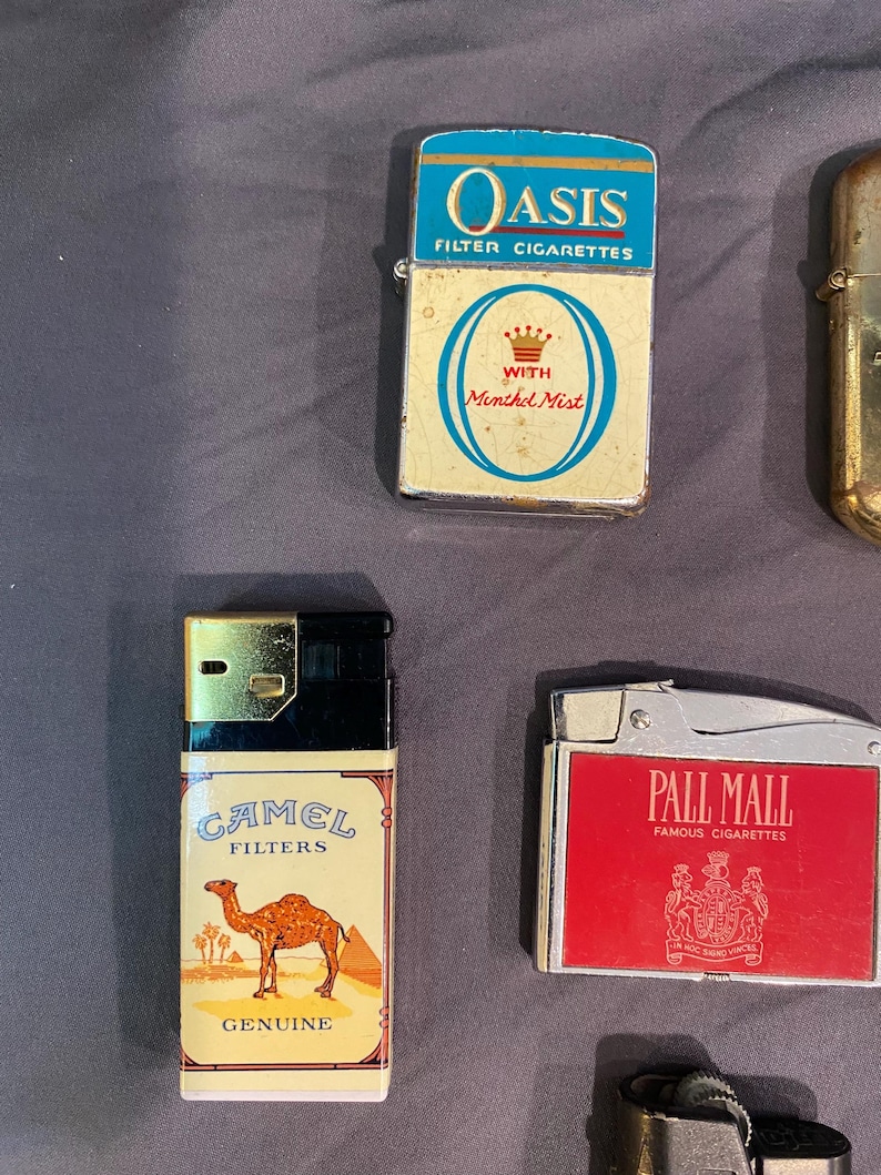 May include: Four vintage cigarette lighters with different brand names. The lighters are silver, gold, and red with various designs. The lighters are all in good condition and have a retro look.