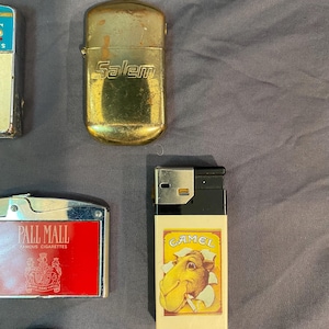 May include: A collection of vintage cigarette lighters. A gold lighter with the word "Salem" on it, a black and white lighter with the word "Camel" on it, a red lighter with the words "Pall Mall Famous Cigarettes" on it, and a black lighter with a silver wheel.