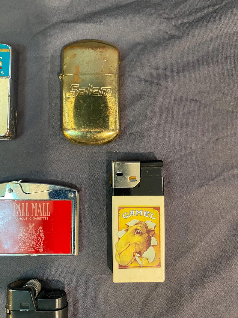 May include: A collection of vintage cigarette lighters. A gold lighter with the word "Salem" on it, a black and white lighter with the word "Camel" on it, a red lighter with the words "Pall Mall Famous Cigarettes" on it, and a black lighter with a silver wheel.
