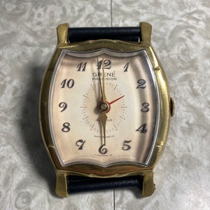 May include: A gold-toned Grene Precision wristwatch with a white face and black leather band. The watch has black Roman numeral hour markers and a red second hand. The watch face also includes the words "Grene Precision" and "Anti-Magnetic".