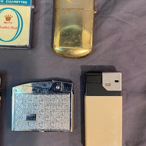 May include: A collection of vintage lighters, including a silver lighter with a crown pattern, a gold lighter, and a black and beige lighter. The lighters are all in good condition and would make a great addition to any collection.