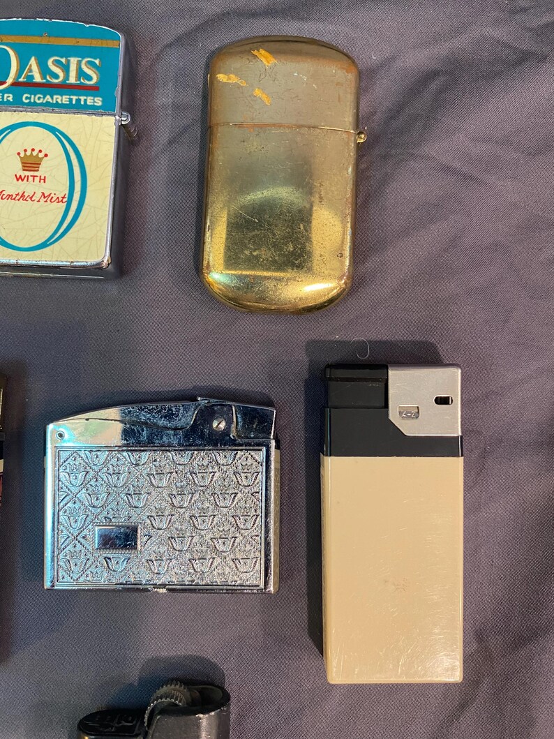 May include: A collection of vintage lighters, including a silver lighter with a crown pattern, a gold lighter, and a black and beige lighter. The lighters are all in good condition and would make a great addition to any collection.