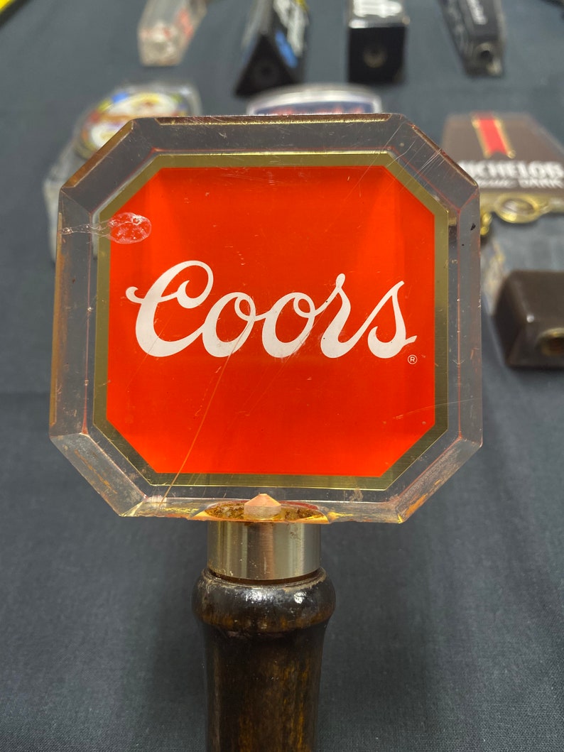 Może przedstawiać: Przezroczysta plastikowa rączka do kranu piwnego z czerwonym tłem i napisem "Coors" białymi literami. Rączka jest przymocowana do drewnianej podstawy.