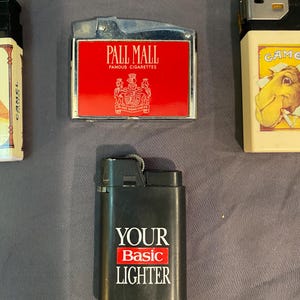May include: A collection of lighters, including a black lighter with the text "YOUR Basic LIGHTER", a red lighter with the text "PALL MALL FAMOUS CIGARETTES", and a white lighter with the text "GAME" and a camel illustration.
