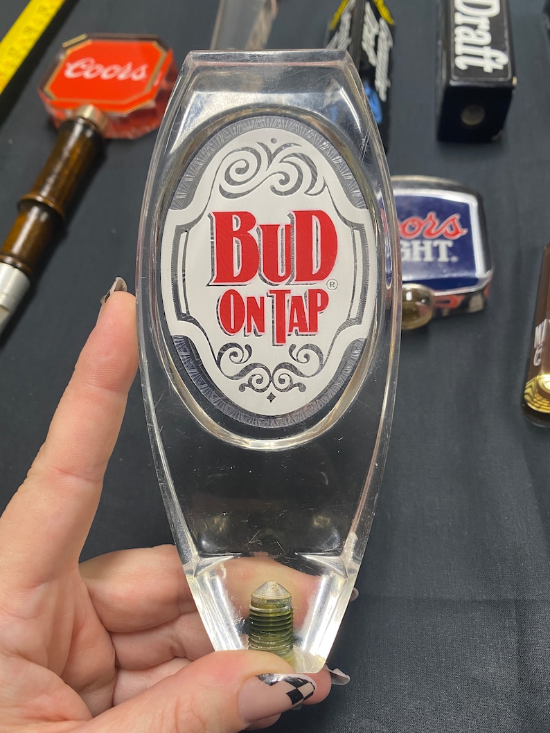 Może przedstawiać: Przezroczysty plastikowy uchwyt do kranu piwnego z białą owalną etykietą z napisem "Bud On Tap" czerwonymi literami.