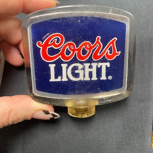Może przedstawiać: Plastikowa, przezroczysta rączka do nalewania piwa Coors Light z niebiesko-białą etykietą i czerwonymi napisami. Rączka ma złotą, gwintowaną podstawę.