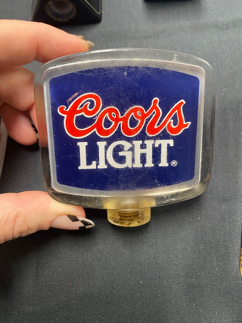 Może przedstawiać: Plastikowa, przezroczysta rączka do nalewania piwa Coors Light z niebiesko-białą etykietą i czerwonymi napisami. Rączka ma złotą, gwintowaną podstawę.