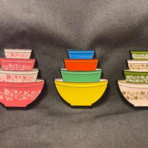 May include: Three sets of nesting mixing bowls, each set with a different color scheme. The first set is pink with white floral designs, the second set is yellow with green, red, and blue bowls, and the third set is white with green floral designs.