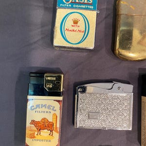 May include: A collection of vintage lighters, including a silver lighter with a patterned design, a black and gold lighter with a Camel Filters Imposter label, a gold lighter, and a blue and white lighter with the Oasis Filter Cigarettes logo and the words "With Menthol Mist".