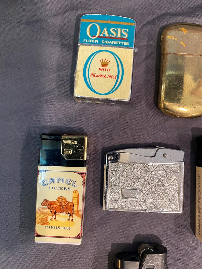 May include: A collection of vintage lighters, including a silver lighter with a patterned design, a black and gold lighter with a Camel Filters Imposter label, a gold lighter, and a blue and white lighter with the Oasis Filter Cigarettes logo and the words "With Menthol Mist".