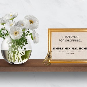 May include: A gold framed print with the text "Thank you for shopping... Simply Minimal Home by Obtaining Abundance Est. 2023" on a beige background. A small gold bird figurine and a glass vase with white flowers are on a wooden shelf.