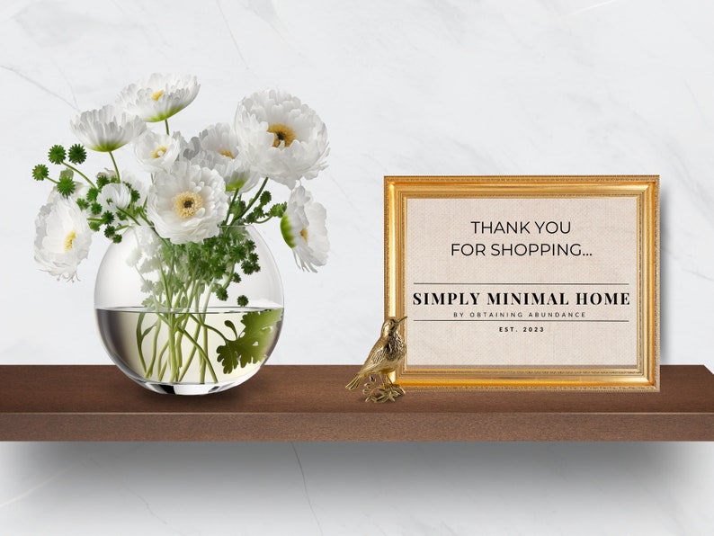 May include: A gold framed print with the text "Thank you for shopping... Simply Minimal Home by Obtaining Abundance Est. 2023" on a beige background. A small gold bird figurine and a glass vase with white flowers are on a wooden shelf.