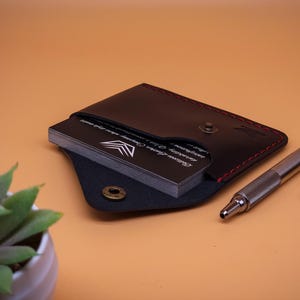 May include: A black leather card holder with red stitching and a brass snap button closure. The holder contains a stack of black business cards with white text and a logo. A silver metal pen and a brown leather watch strap are positioned nearby.