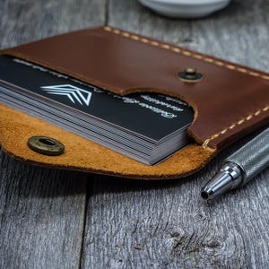 May include: A brown leather card holder with a snap button closure containing a stack of black business cards with a white chevron logo and text. A silver metallic pen with a textured grip is positioned next to the card holder.