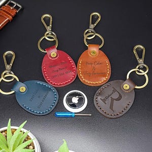May include: Four oval leather keychains in blue, red, tan, and dark brown with brass hardware, lobster clasps, and stitching. The keychains feature engraved text including "World's Best Dog Ever!!", "Keep Calm & Stay Awesome", and the letter "R".
