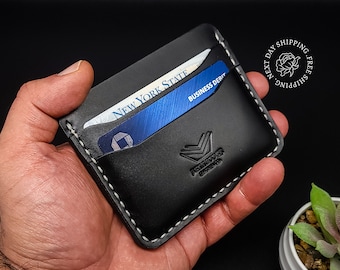 Puede incluir: Cartera de cuero negro con costuras blancas y un logotipo que dice "Asymmetrical Designs". La cartera tiene dos ranuras para tarjetas y una ranura para tarjetas de visita. La cartera se muestra con tarjetas en su interior.