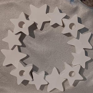 May include: A white star-shaped wreath with a circular center. The wreath is composed of multiple star-shaped pieces, some with a circular hole in the center. The stars are arranged in a circular pattern.