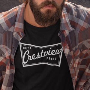 May include: A black t-shirt with the text "Crestview Pride" in a white, stylized font. The shirt is worn with a plaid button-down shirt. The person in the image has a beard and long hair.