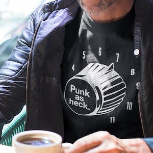 May include: A black t-shirt with the text "Punk as heck." and a graphic of a speaker. The shirt is worn with a black leather jacket. A white coffee cup and saucer are on a wooden table.