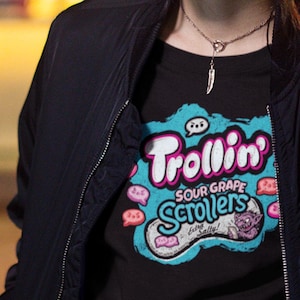 May include: Black t-shirt with a colorful graphic that reads "Trollin' Sour Grape Scrollers". The design features a cartoonish character and speech bubbles. The person is wearing a black jacket.