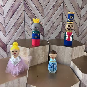 May include: Hand-painted wooden peg dolls, including a mouse with a crown, a soldier, a ballerina, and a girl in a blue dress. The dolls are displayed on hexagonal platforms against a wood-paneled backdrop. These dolls are perfect for collectors.