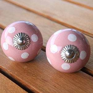 May include: Two pink ceramic knobs with white polka dots and silver accents. The knobs are round and have a shiny finish.