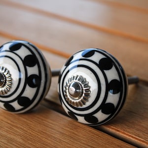 May include: Two black and white ceramic cabinet knobs with a silver center. The knobs have a geometric pattern with black circles and white lines.