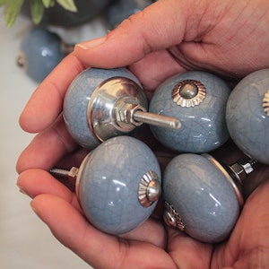 Puede incluir: Una colección de seis pomos de armario de cerámica gris con detalles plateados. Los pomos tienen un acabado craquelado y se sostienen en una mano.