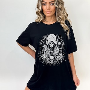 May include: Black t-shirt featuring a white graphic print of a hooded figure with horns, holding a skull. The figure is surrounded by stars and a crescent moon.