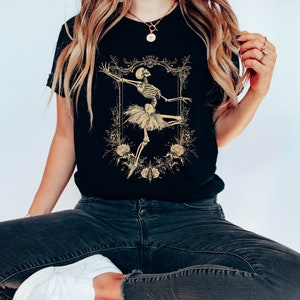 Może przedstawiać: Czarny T-shirt z grafiką szkieletu ubranego w baletnicę w złotej ramie z kwiatowymi akcentami. Tekst "Forever" znajduje się nad ramą.