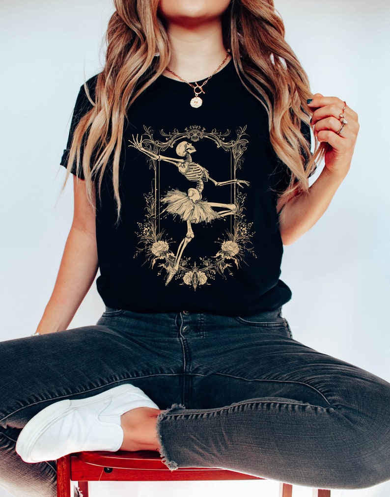 Może przedstawiać: Czarny T-shirt z grafiką szkieletu ubranego w baletnicę w złotej ramie z kwiatowymi akcentami. Tekst "Forever" znajduje się nad ramą.