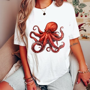 May include: White t-shirt with a red octopus graphic. The octopus has eight tentacles and a large eye. The shirt is being worn by a person.