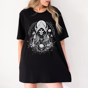 May include: Black t-shirt featuring a white graphic of a hooded figure with horns, a skull, and a moon in a woodland setting.