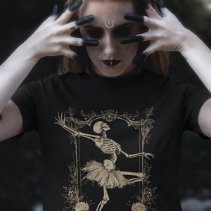 Może przedstawiać: Czarny T-shirt z grafiką złotego szkieletu baletnicy. Szkielet ma na sobie tutu i jest otoczony kwiatowymi detalami.