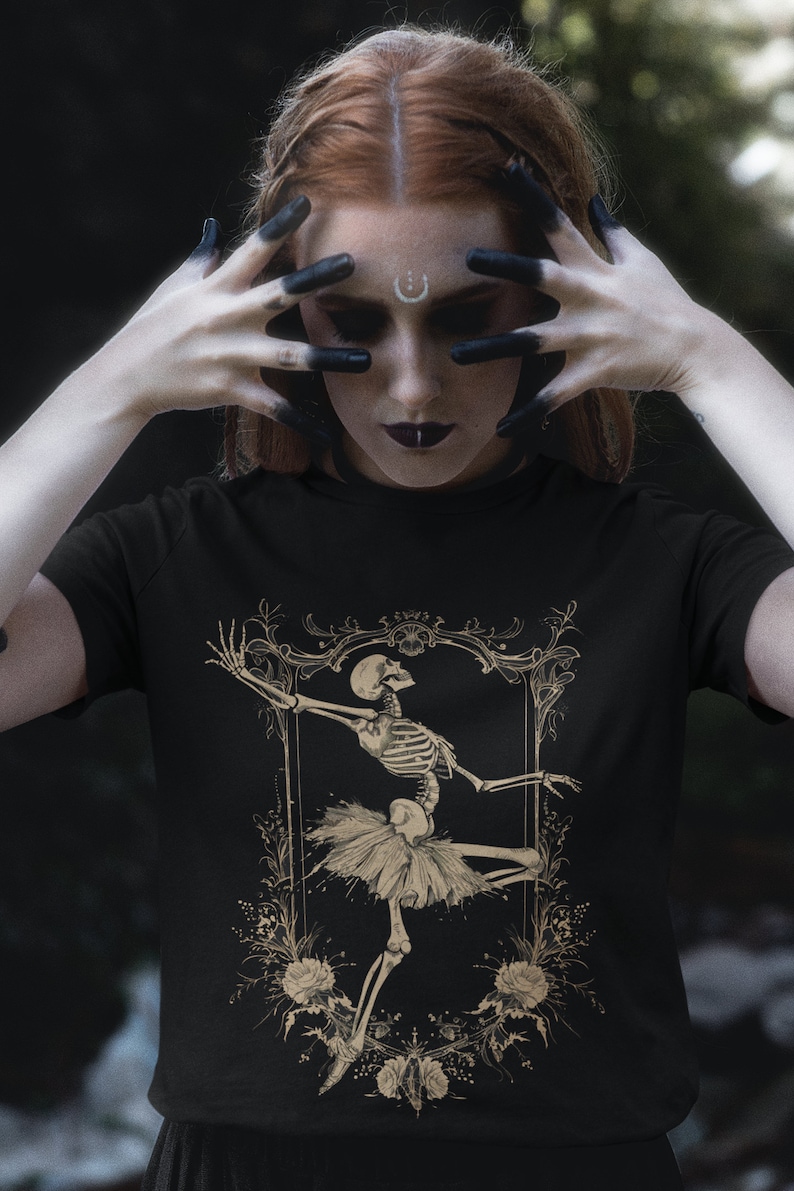 Może przedstawiać: Czarny T-shirt z grafiką złotego szkieletu baletnicy. Szkielet ma na sobie tutu i jest otoczony kwiatowymi detalami.
