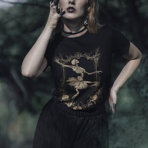 Może przedstawiać: Kobieta w czarnym t-shircie z nadrukiem złotego szkieletu baletnicy. Koszulka ma vintage'owy, gotycki styl. Ma na sobie czarną sp&oacute;dnicę i czarne paznokcie.
