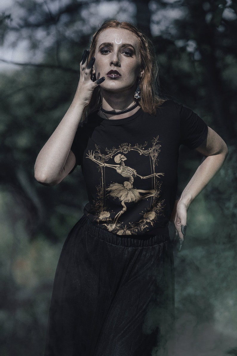 Może przedstawiać: Kobieta w czarnym t-shircie z nadrukiem złotego szkieletu baletnicy. Koszulka ma vintage'owy, gotycki styl. Ma na sobie czarną sp&oacute;dnicę i czarne paznokcie.