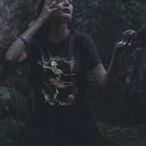 Może przedstawiać: Kobieta w czarnym t-shircie z nadrukiem złotej baletnicy-szkieletu. Trzyma w prawej ręce kryształową kulę, a lewą rękę ma na czole. Tło stanowi ciemny las.