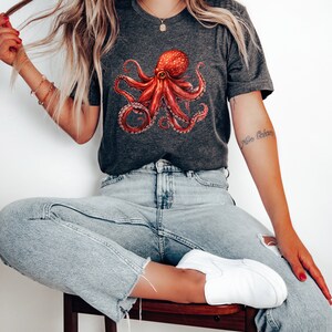 May include: A dark gray t-shirt with a red octopus graphic. The octopus has eight tentacles and is in a relaxed pose. The shirt has a crew neck and short sleeves.