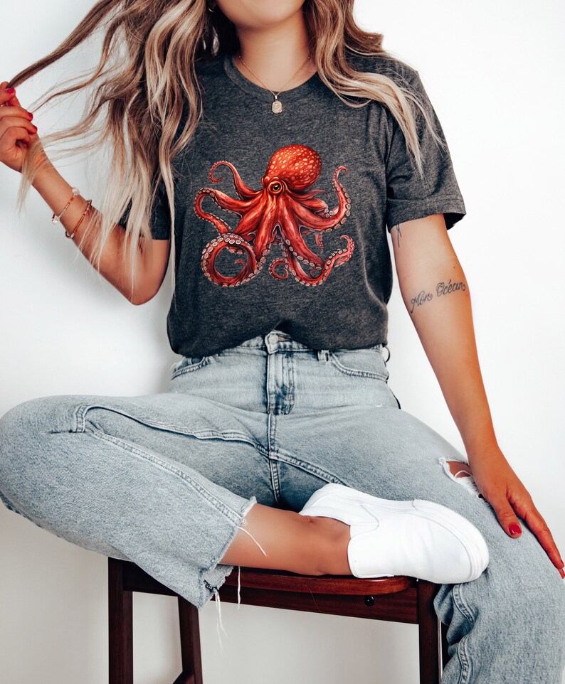 May include: A dark gray t-shirt with a red octopus graphic. The octopus has eight tentacles and is in a relaxed pose. The shirt has a crew neck and short sleeves.