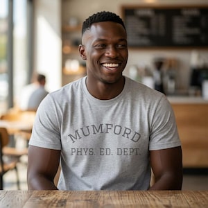 Peut inclure: Un t-shirt à manches courtes et col rond gris clair avec le texte gris foncé MUMFORD arqué au-dessus des mots PHYS. ED. DEPT. imprimés dans une police universitaire classique sur la poitrine du vêtement.