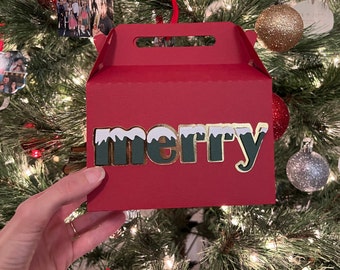 May include: Red gift box with a white handle and the word "merry" in gold letters with green and white accents. The box is sitting on a Christmas tree.