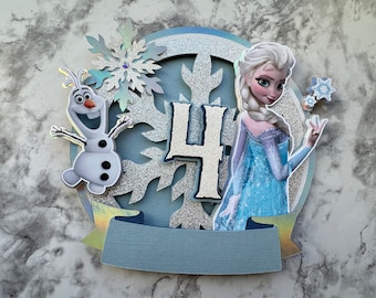 May include: A blue and silver cake topper featuring Elsa from Frozen, Olaf, and the number 4. The topper is decorated with snowflakes and glitter.