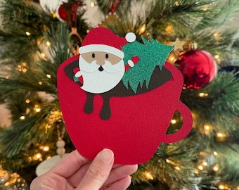 May include: A red paper cup with a brown rim and a green paper Christmas tree inside. A paper Santa Claus with a white beard is sitting in the cup.