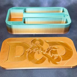 May include: A two-toned, 3D-printed storage box with a lid. The box is teal and gold, with a rectangular shape and multiple compartments. The lid is gold and features the letters "D&D" with a dragon graphic.