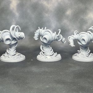 May include: Three gray miniature tornado figurines. Each figurine has a swirling, textured design, with a circular base. The figurines are arranged in a row, with the middle one slightly taller than the others. The background is a mottled gray.