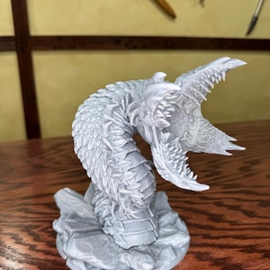 May include: A gray, detailed sculpture of a serpent with an open mouth, revealing sharp teeth. The serpent is coiled and rests on a rocky base. The sculpture has a textured, scaly appearance, suggesting a fantasy or mythological theme.
