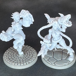 May include: Two light gray miniature figurines of female characters. One figure is posed with a large mechanical gauntlet, the other with a shark-shaped weapon. Both figures are on textured circular and polygonal bases.
